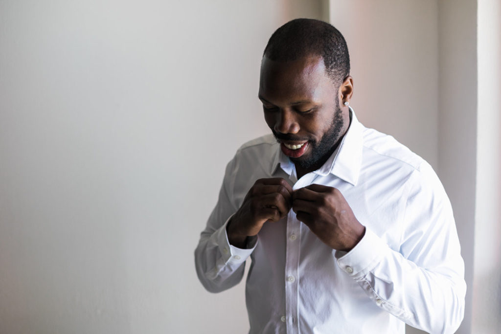 Groom smiles as he fastens his shirt before his wedding