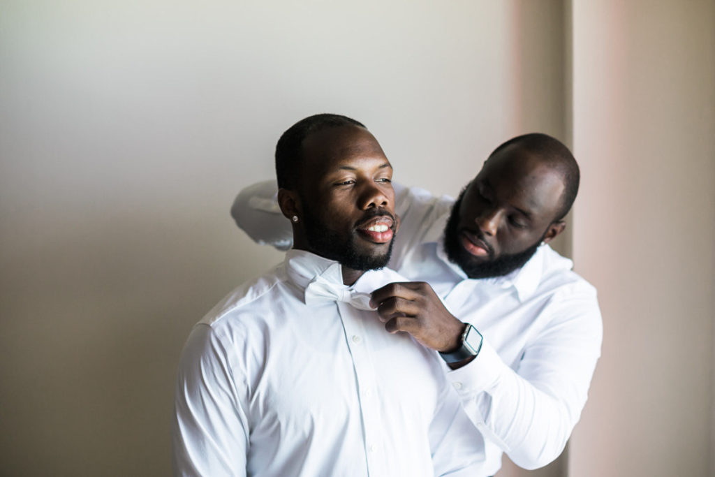 Groomsman fastens bow tie on groom as he looks out the window