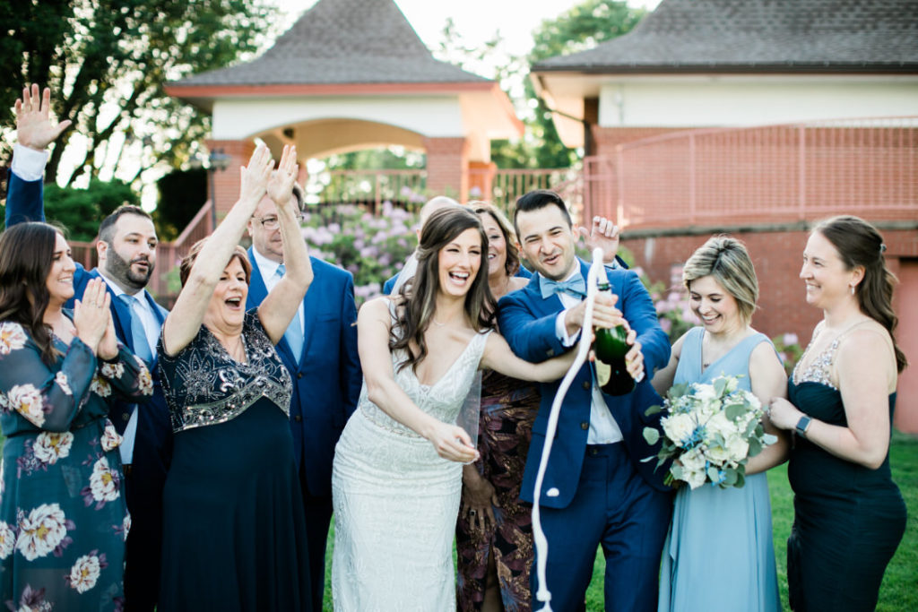 Group of family and friends cheer as groom and bride pop bottle of champagne