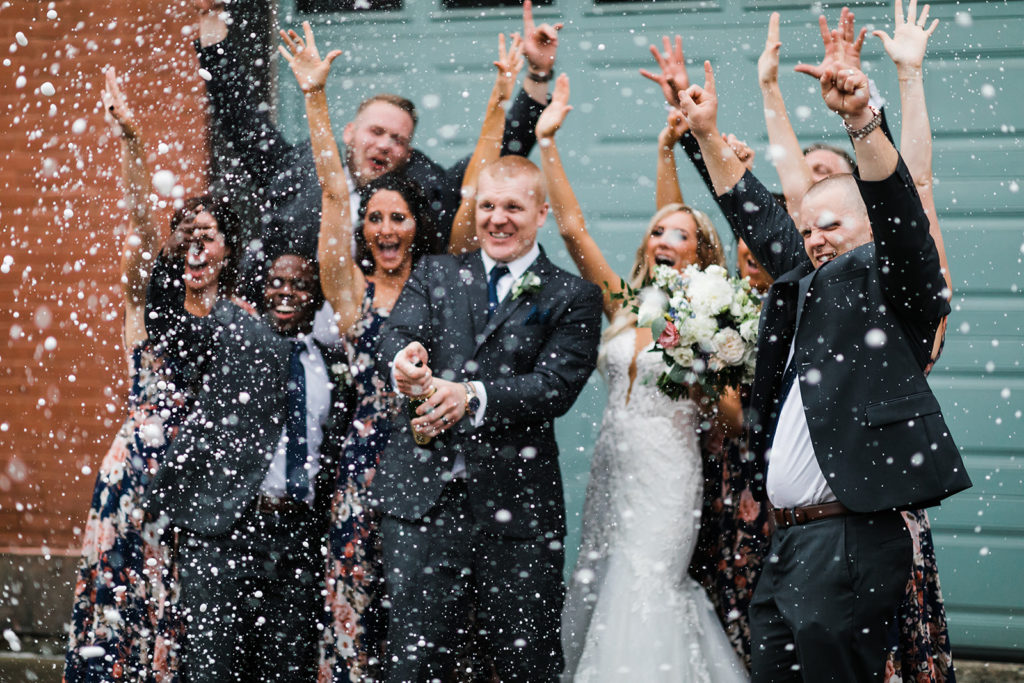 Bridal party cheers as groom pops a champagne bottle