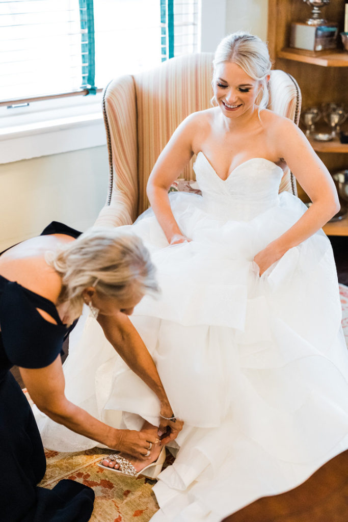 bride smiles as she sits and her mother straps her wedding shoes