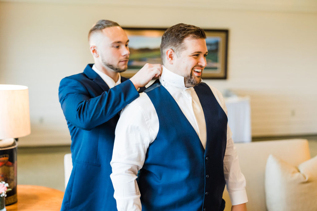 Groom smiles as a groomsman adjusts the collar of his shirt