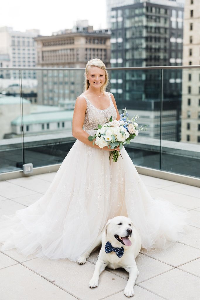 Bride smiles and holds her bouquet with her dog laying at her feet