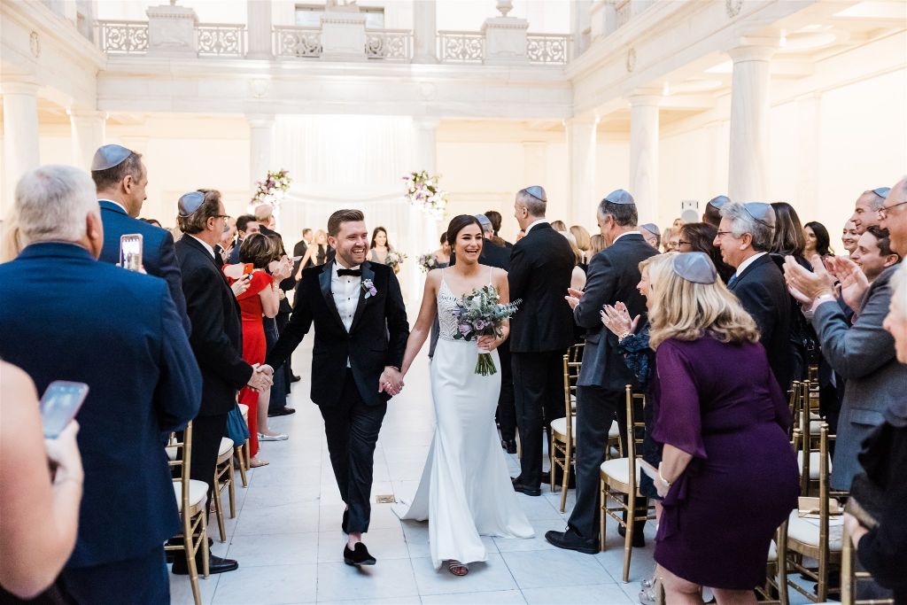 bride and groom smile and walk up the aisle at Jewish wedding at the Carnegie