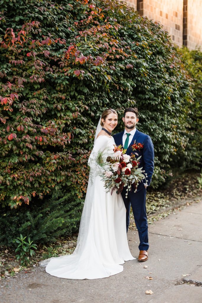 Bride and groom pose together against wall of fall greenery