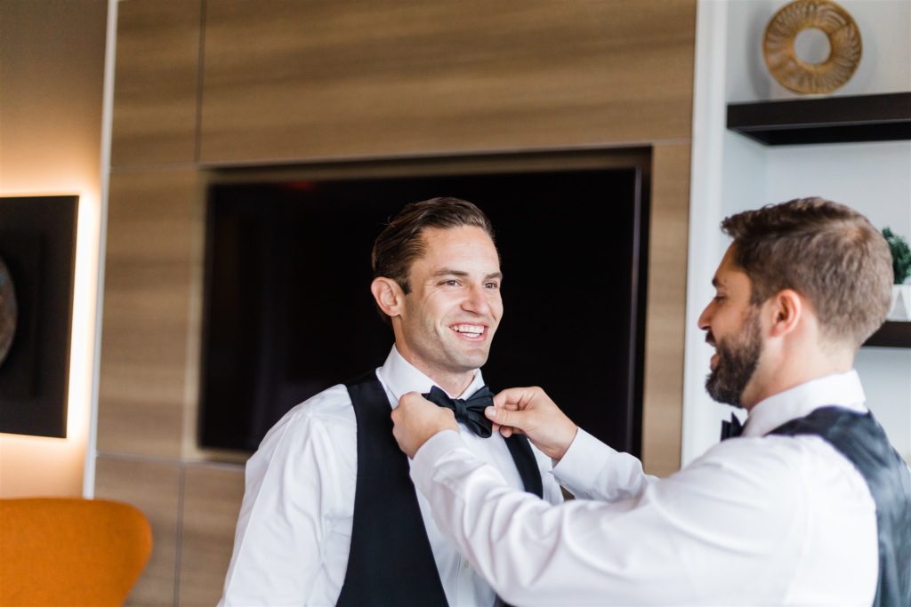 Groom smiles as groomsman adjusts his bow tie