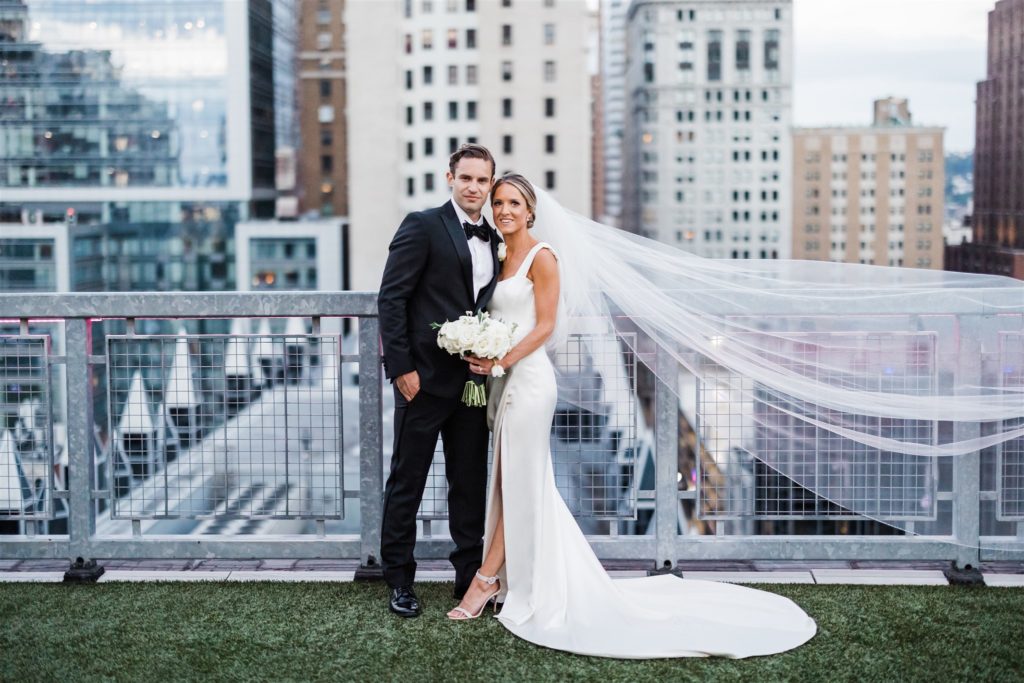 Bride and groom smile into the camera from a rooftop view over Pittsburgh